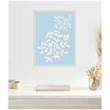 Framed pale blue canvas print featuring white botanical silhouettes of leaves and flowers, displayed on a white wall above a light wood and white desk with dried flower decor.