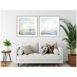 Two framed watercolor prints above a white sofa, depicting soft pastel skies and seascapes with pink, blue, and beige hues, flanked by leafy green plants.
