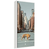 Framed vertical canvas showing a tiger walking across a city street lined with tall buildings under a clear blue sky.
