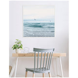 Square framed canvas depicting a serene ocean scene with gentle waves under a pale sky and two birds flying, hung above a minimalist wooden desk and gray chair.