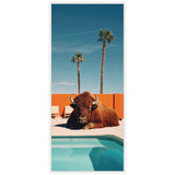 Large brown bison resting on poolside concrete with clear blue water and an orange wall behind, two tall palm trees under a bright blue sky.