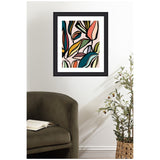 Framed abstract botanical print featuring colorful stylized ficus leaves in muted pink, coral, teal, green, and yellow, hanging on a white wall above an olive-green chair and potted plant.