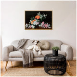 Gray sofa with a textured throw and round pillow, a black rattan side table holding a small plant, and a framed floral canvas with orange, pink, and white flowers on a dark background above.