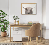 Minimalist office nook with light wood desk, beige upholstered chair, green potted plant, and framed Highland cow portrait with soft gray and cream tones.