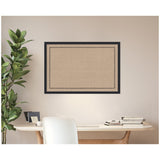 Rectangular cork pinboard framed in black with a subtle scallop pattern border, mounted on a beige wall above a wooden desk with books and a plant.