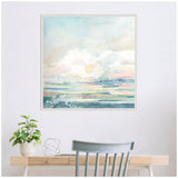 Abstract watercolor landscape painting with soft pastel clouds and layered horizontal bands of green, pink, and blue, framed with a white textured border, hanging above a light wood desk.