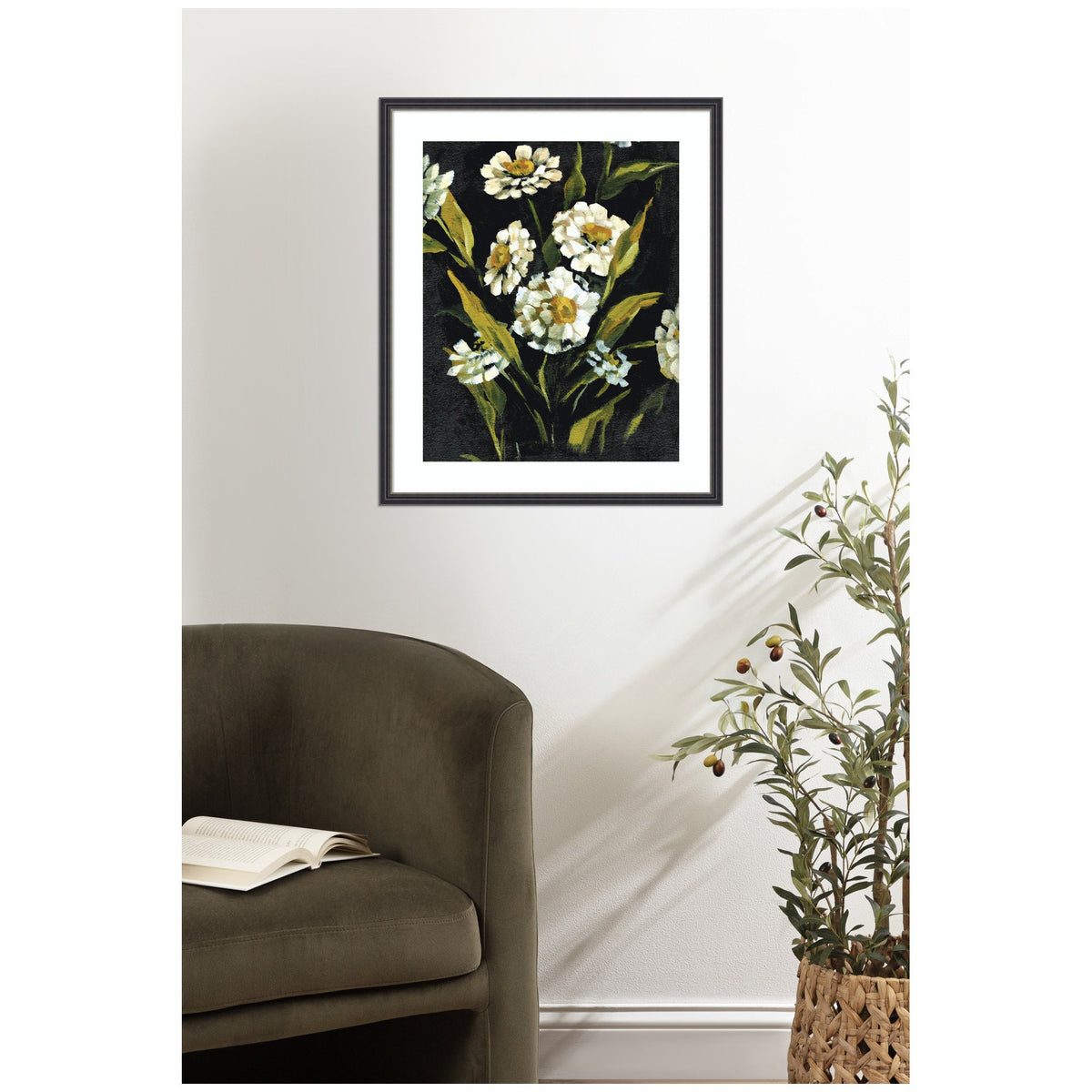 Framed painting of white flowers with green leaves on a dark background, hanging above an olive green curved armchair and a plant in a woven basket.