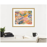 Abstract floral painting with overlapping leaves and petals in warm pink, orange, green, and blue tones, framed in light wood above a desk with a book, plant, and coffee cup.