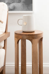 Round wooden drink table with curved legs and natural wood grain, holding a white ceramic mug with a tea bag tag hanging from the rim, positioned next to a light-colored fabric chair.