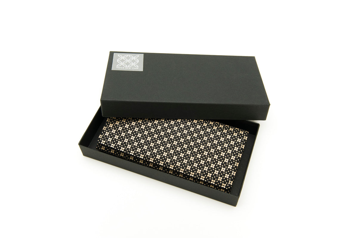 Black long wallet with a cream flower lattice pattern resting in a black box with a silver decorative label on the lid corner, shown from an angled top view.