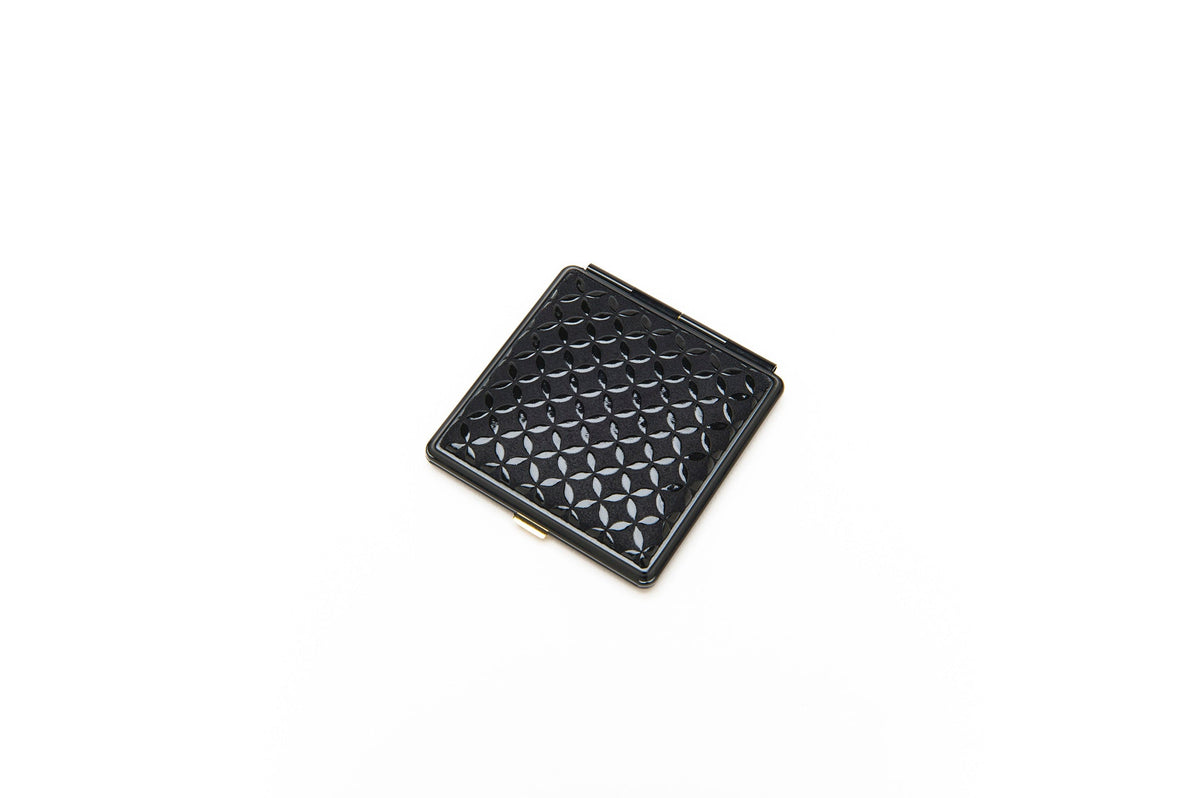 Square black compact mirror featuring a textured geometric pattern of interlocking circles on its surface, shown closed on a white background.