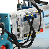Close-up of a transparent plastic control box with blue internal components and multiple black braided cables connected, mounted on a metallic blue CNC router frame with a red power switch visible.