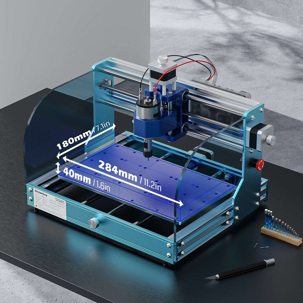 Compact desktop CNC router with teal frame, blue protective shields, and labeled working area dimensions, placed on a black surface next to a precision tool set and pen.