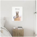 Framed canvas of a raccoon sitting in a wicker basket with colorful laundry, titled 'little rascal laundry service,' hanging above a wooden bedside table with books and a cup.