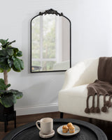 Black metal arched wall mirror with ornate floral detailing at the top, reflecting a window; shown above a black tray table holding a mug and three muffins, beside a white boucle armchair with a brown tasseled throw.