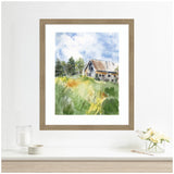 Watercolor painting of a weathered barn amidst green grass and trees under a bright blue sky, framed in light wood and mounted on a white wall.