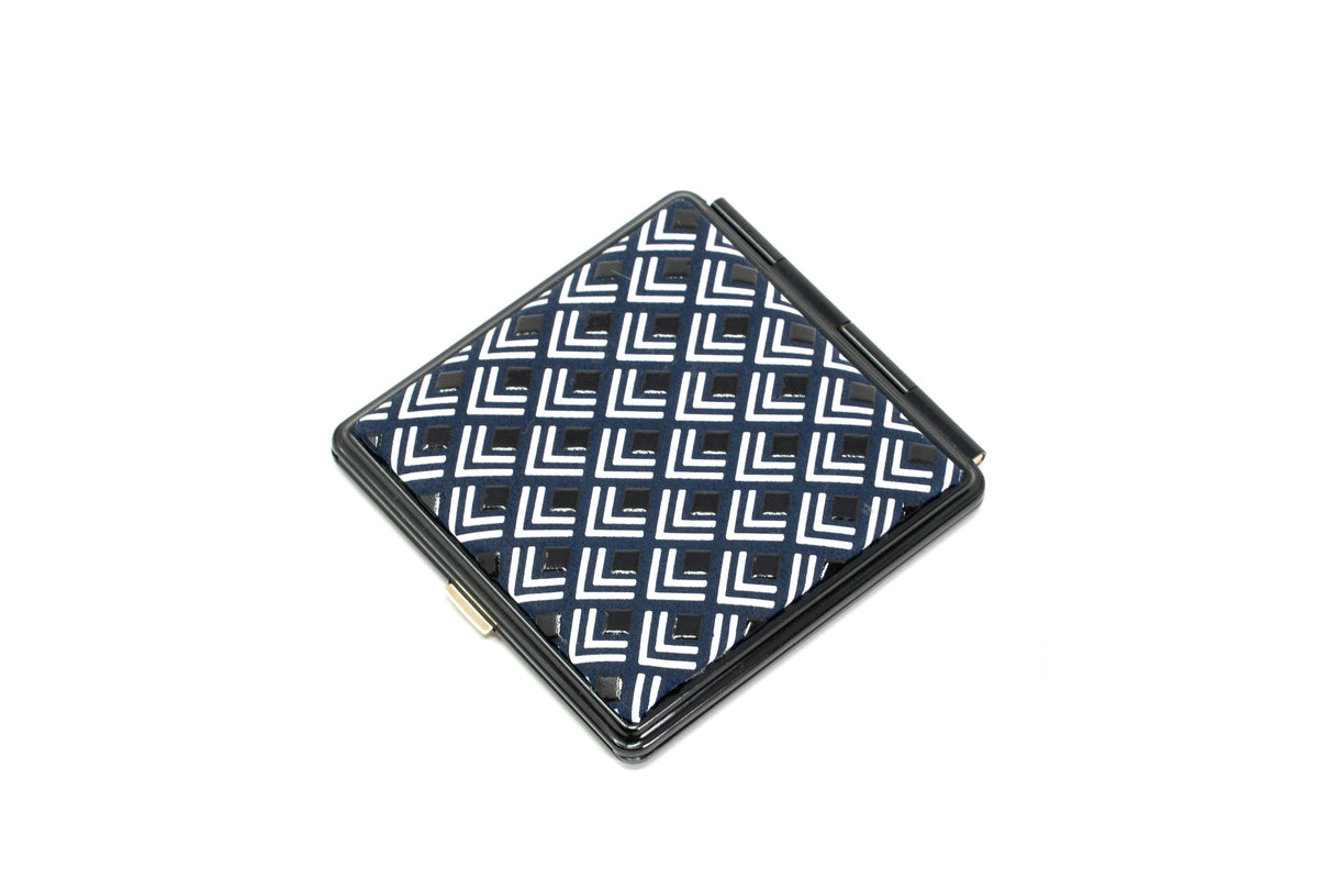 Square compact mirror with a repeating pattern of white and blue geometric L shapes on the top surface against a black frame.
