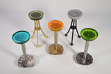Five round Martini Tables with translucent resin tops in green, orange, gray, and blue, each featuring unique imprinted patterns and supported by metal legs in silver, gold, and black.