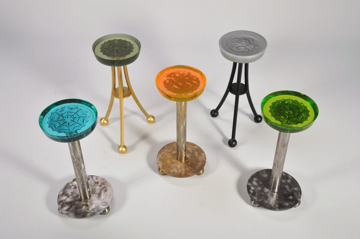Five round Martini Tables with translucent resin tops in green, orange, gray, and blue, each featuring unique imprinted patterns and supported by metal legs in silver, gold, and black.