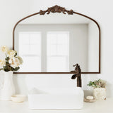 Arched mirror with an ornate bronze frame and decorative crest above a white rectangular vessel sink, flanked by a white vase with cream roses and a small floral planter on a bathroom countertop.