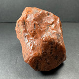 Rough reddish-brown mahogany obsidian rock with glossy and matte textured surfaces, irregular shape, and dark speckles, resting on a dark gray background.