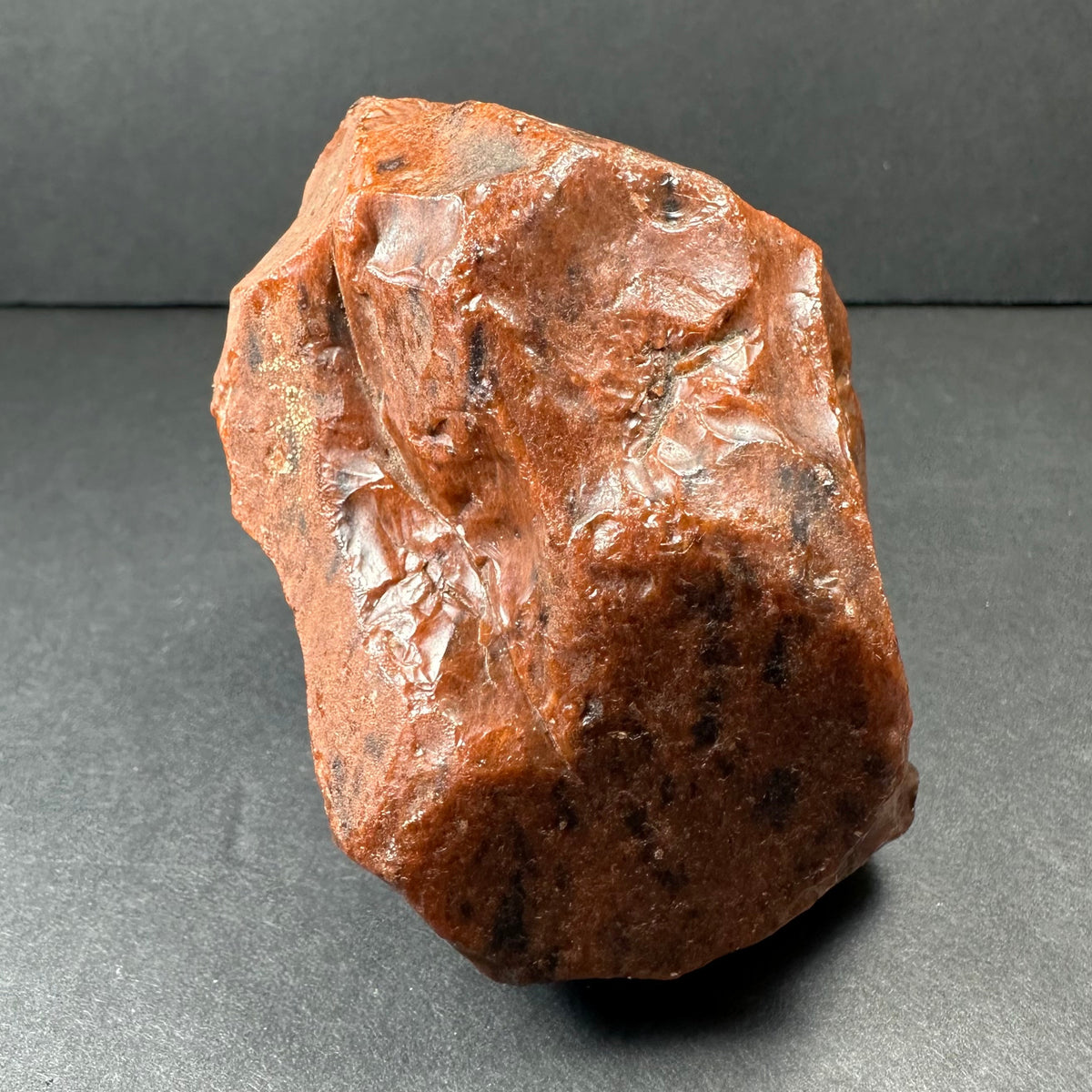 Rough reddish-brown mahogany obsidian rock with glossy and matte textured surfaces, irregular shape, and dark speckles, resting on a dark gray background.