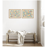 Two framed canvases with rows of multicolored dots on a white background, hung side by side above a wooden bench with a draped beige blanket and open book.