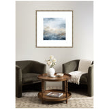 Abstract framed wall art with misty blue and beige tones hanging above two dark green armchairs and a round wooden table with white vase and decor, on a patterned rug.