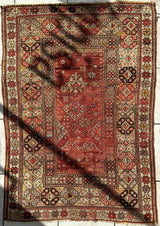 Rectangular antique Turkish wool prayer rug with distressed red central field, geometric floral motifs, and multi-layered beige, brown, and red patterned borders.