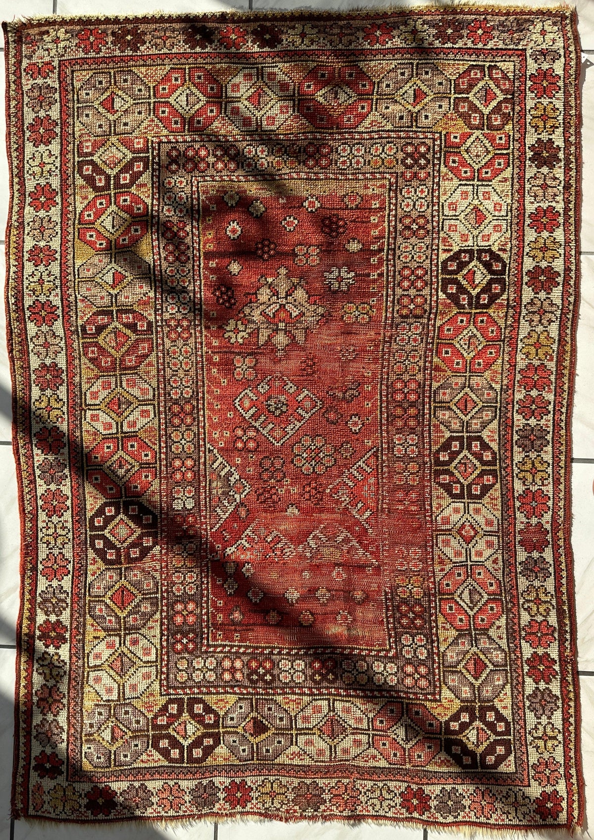 Rectangular antique Turkish wool prayer rug with distressed red central field, geometric floral motifs, and multi-layered beige, brown, and red patterned borders.