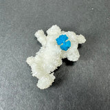 Small white stilbite specimen with clustered crystal formations and a vivid blue cavansite ball resting on top, displayed on a dark gray surface.