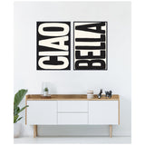 Two black and white framed canvases with bold uppercase typography reading 'CIAO' and 'BELLA' hung side by side above a minimalist white and wood sideboard with decorative items.
