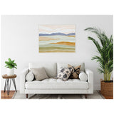 Framed canvas of soft pastel hills in beige, blue, and green above white sofa with patterned pillows, flanked by potted plants in a minimal living room.