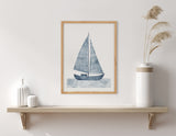 Framed minimalist print of a blue sailboat with striped sails above stylized wavy water, displayed on a light wood shelf with white and beige pottery and dried grass.
