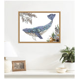 Framed canvas print of a blue whale composed of overlapping blue and purple leaf shapes, surrounded by detailed botanical elements, hanging above a white console with a lamp, photo frame, and potted plant.