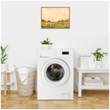 Framed canvas print above a white front-loading washing machine showing cows grazing in a grassy field under a warm, orange sky, hung on a white wall in a laundry room.