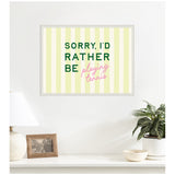 Framed wall art with vertical pale yellow stripes and text saying 'SORRY, I'D RATHER BE playing tennis' in dark green and pink script above a white table with lamp, photo, and plant