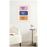 Three vertically stacked vintage-style tickets in pink, navy blue, and yellow with "LUCKY YOU" text and star accents framed on a white wall above a small round table and cream chairs.