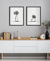 Two minimalist black and white botanical sketches in black frames showing a tall bottle with small flowers and a round vase with clustered flowers, hung above a modern white sideboard with wood top.