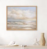 Framed artwork depicting a calm seascape with gentle beach waves under soft, cloudy sunset skies in pale blues and warm beige tones, displayed on a white wall above a natural wooden shelf with coastal decor.