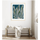 Framed abstract botanical art with elongated leaf shapes in cream, green, and blue on a deep teal background, hanging above a rustic wooden bench with a draped white cloth.