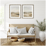 Two framed abstract landscape prints in soft beige and gray tones hung above a wooden bench with white and gray checkered pillows, flanked by woven baskets and a large potted plant.