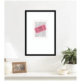 Framed artwork of a pink ticket labeled ONE LIFE placed over a newspaper page titled The Good News, hanging on a white wall above a table with lamp, photo frame, and potted plant.