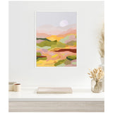Framed canvas print of abstract landscape with pastel hills in pink, green, and yellow tones under a pale sky with a sun, hanging above a white desk with neutral decor.