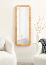Tall rectangular wall mirror with rounded corners and a textured light wooden frame, mounted on a white wall between two off-white armchairs with simple cushions and a white side table with a vase of green branches.