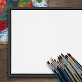 Square blank white canvas in a dark wood frame with a collection of paintbrushes arranged at the bottom right corner on a wooden surface next to a colorful artist palette.
