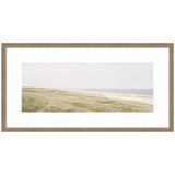 Wide coastal landscape showing sandy beach with gentle waves and grassy dunes under a pale sky, framed in light wood with white mat board.