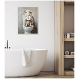 Framed canvas art of a fluffy sheep sitting on a toilet, holding a roll of toilet paper in its mouth, displayed on a white bathroom wall above a freestanding tub.