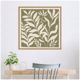 Framed botanical art with off-white leaf silhouettes on olive green canvas above a light wood desk with a potted plant and a minimalist decor piece.