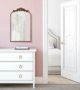 Arched mirror with ornate bronze frame hanging on pink wall above white dresser with gold ring pulls, next to an open door revealing a white bed with patterned bedding.